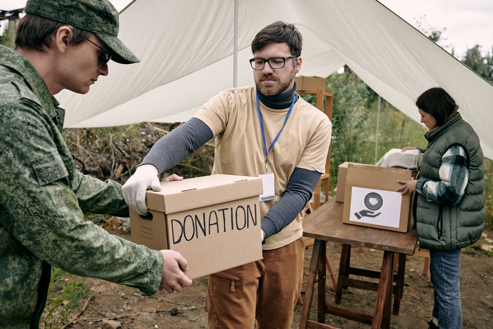 A man hands a donation box to a person in military uniform at an outdoor aid station, whilst another person sorts boxes at a table in the background.