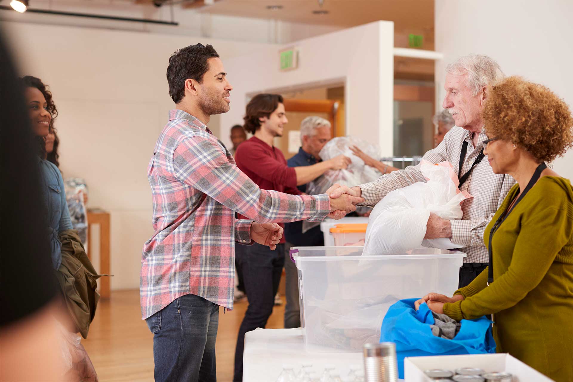 People at a donation centre; a man in a checked shirt shakes hands with a volunteer across a table filled with bags and containers. Other volunteers assist in the background.