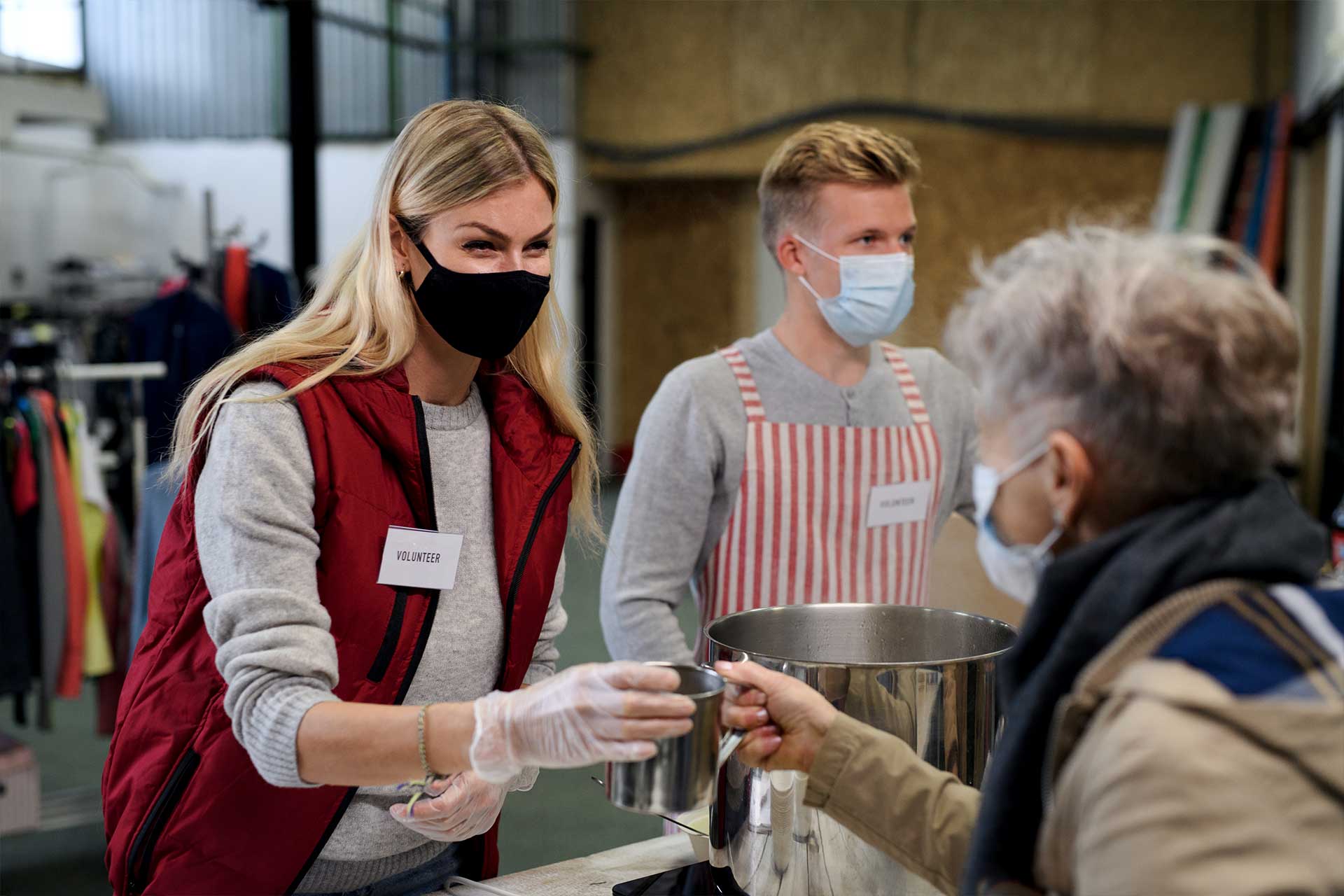 Two volunteers wearing masks serve a meal to an elderly person at a soup kitchen. One volunteer hands a cup to the person, both volunteers wear name badges and gloves.