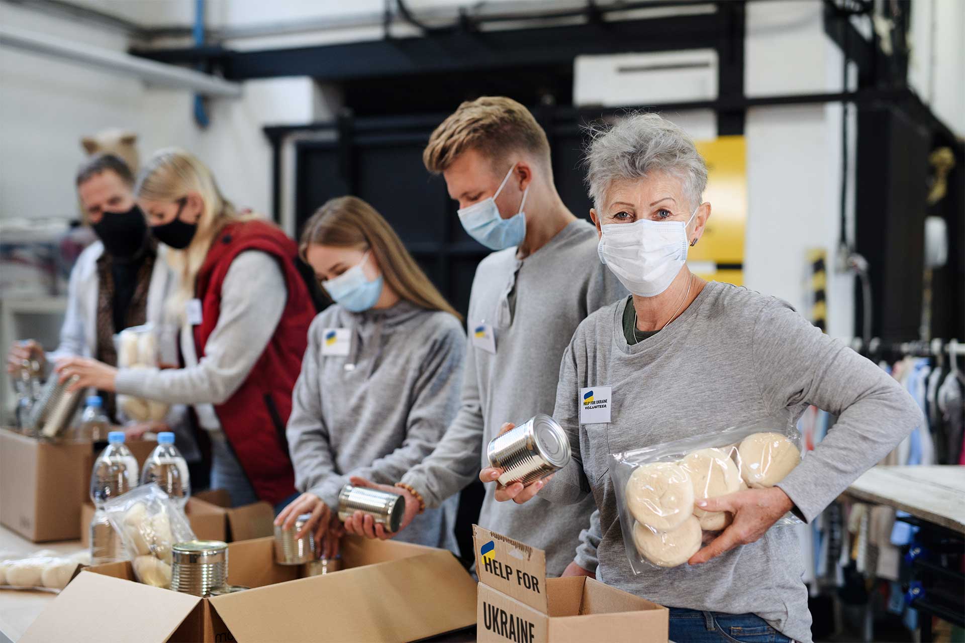 A group of people wearing masks pack food and supplies into boxes labelled "Help for Ukraine" in an indoor setting.