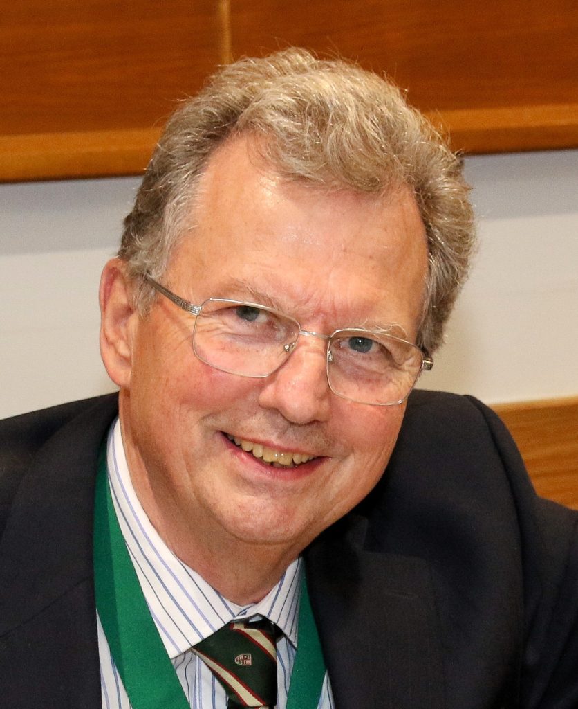 An older man with grey hair and glasses smiles at the camera, wearing a dark suit, striped shirt, and green lanyard.